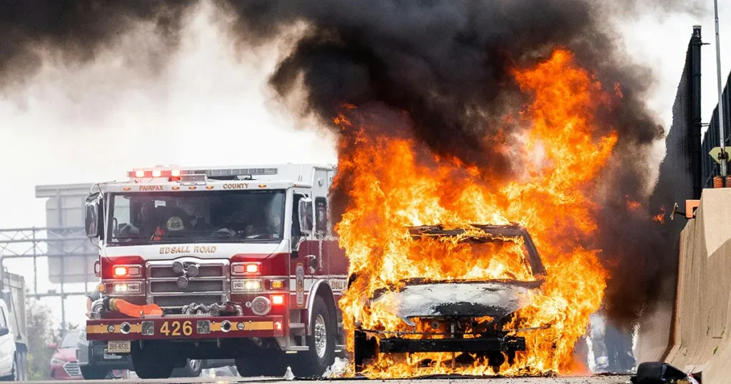 Car engulfed in flames after a car accident with a fire truck to the left of it on a highway.